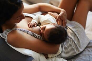 serene loving young black mother in nightgown sitting on bed and breastfeeding baby in comfortable position