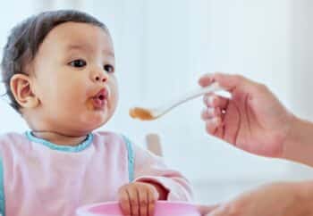 mom feeding baby girl baby food in highchair