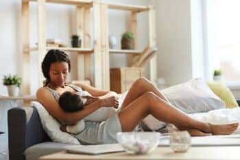 young black mom sitting on sofa bed and breastfeeding baby while holding son in arms