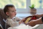 Mother gives toddler baby fruits and berries on a plate. Surprised child takes food from mom woman hands. Kid aged one year and two months