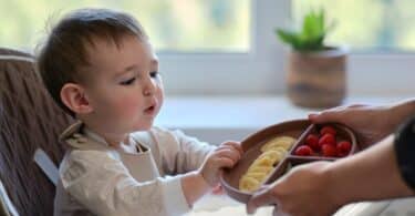 Mother gives toddler baby fruits and berries on a plate. Surprised child takes food from mom woman hands. Kid aged one year and two months