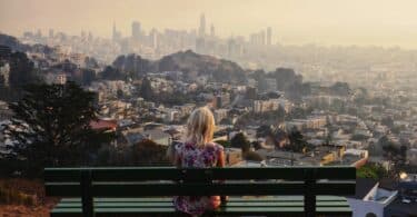 View Of Girl Looking At Cityscape While Sitting On Bench Against Clear Sky