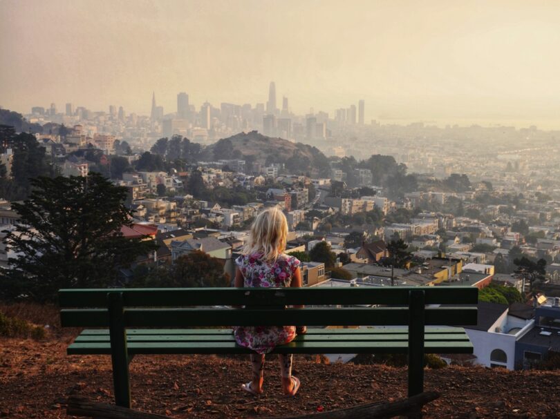 View Of Girl Looking At Cityscape While Sitting On Bench Against Clear Sky